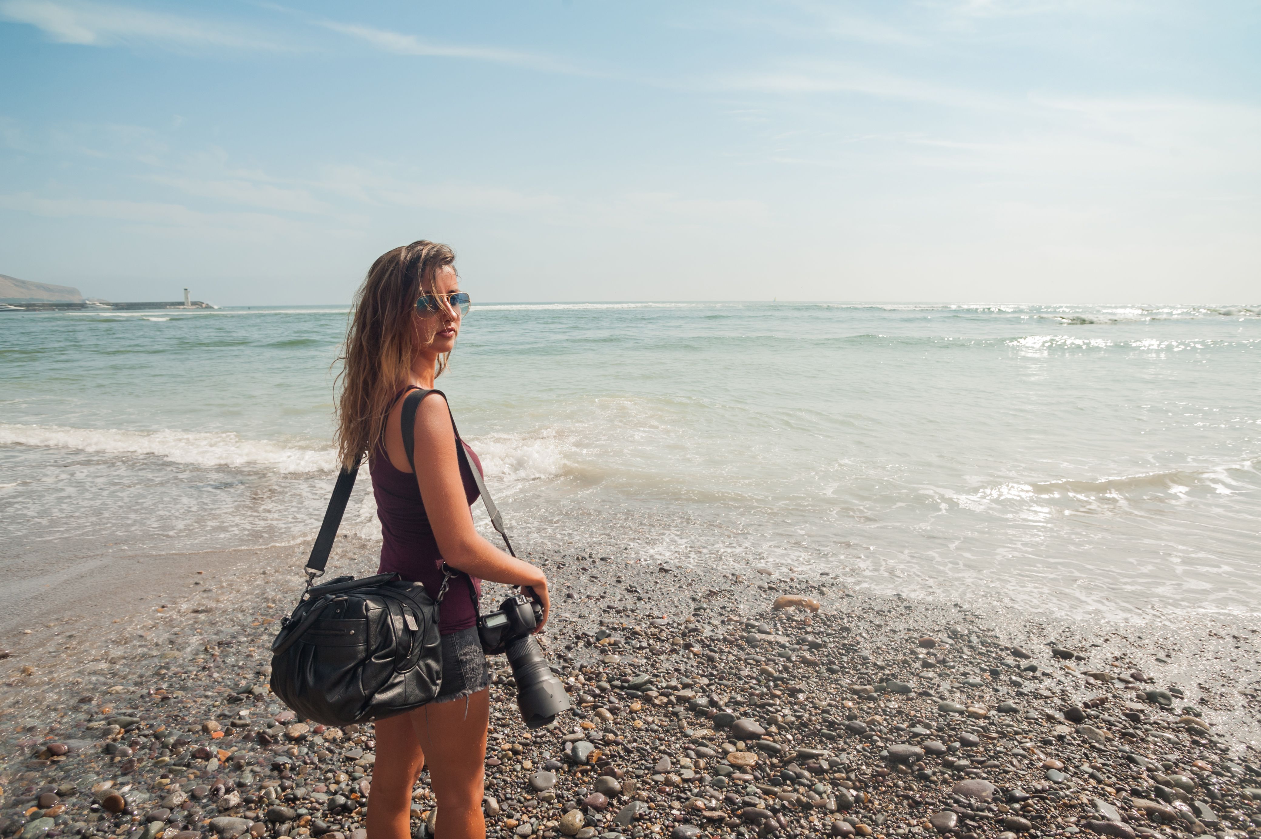 Young woman photographer enjoying peaceful seaside vacation, standing on the beach by the ocean on a sunny day.