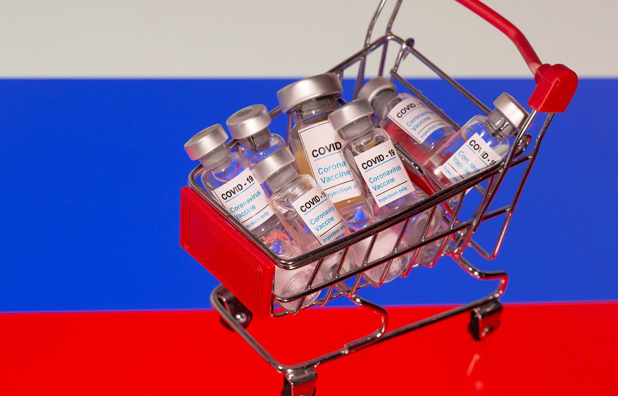 A small shopping basket filled with vials labeled  "COVID-19 - Coronavirus Vaccine"  is placed on a Russian flag