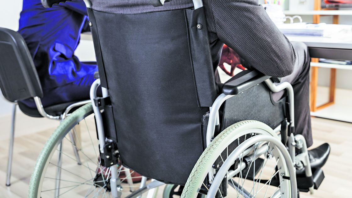 Rear View Of Businessman Sitting In Wheelchair With Colleague At Desk In Office