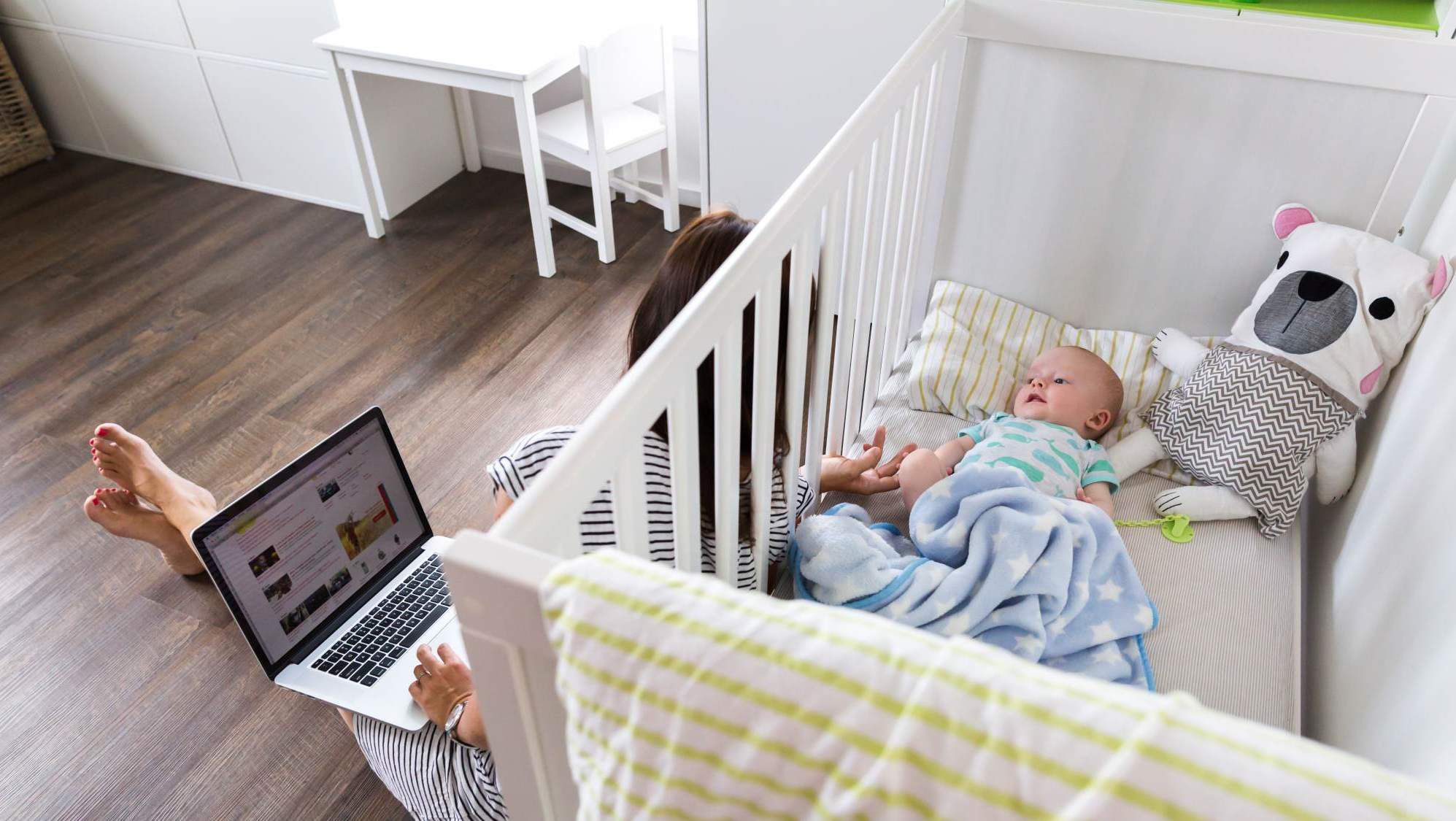 pv-lg-mom-working-on-a-laptop-and-comforting-her-baby-in-a-crib-default-stock-photo-e1558994669680.jpg